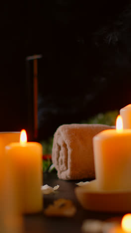 Vertical-Video-Still-Life-Of-Lit-Candles-With-Scattered-Petals-Incense-Stick-And-Soft-Towels-Against-Dark-Background-As-Part-Of-Relaxing-Spa-Day-Decor